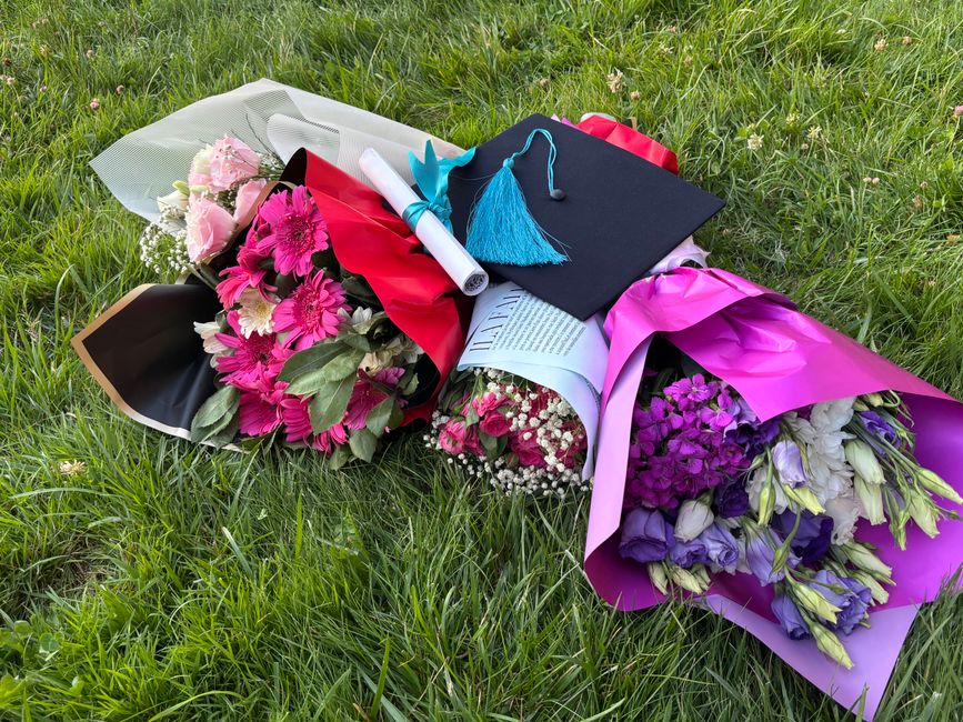 A collection of colorful floral arrangements and bouquets lie on lush green grass, including pink gerbera daisies, purple and white lisianthus, and baby's breath, all wrapped in vibrant pink and purpl