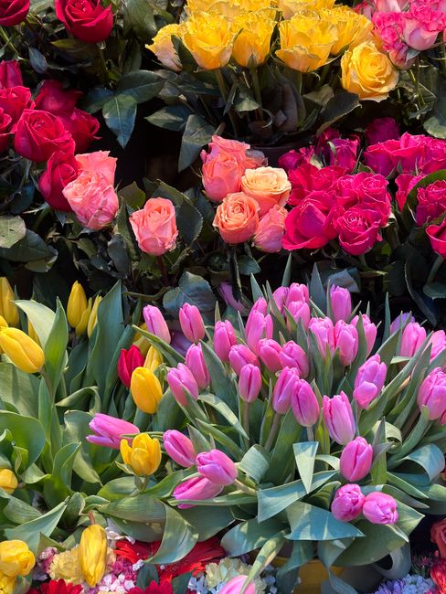 A vibrant floral display featuring a variety of fresh flowers, including pink and yellow tulips with sleek green leaves, and clusters of pink, yellow, and orange roses arranged in a lush, colorful bou