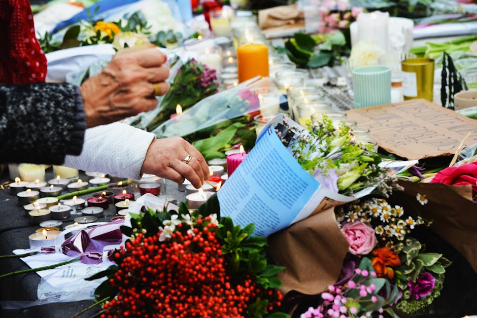 A close-up of a hand placing a small candle next to a floral tribute, with a variety of fresh flowers including pink roses, white daisies, and red berries, arranged on a table covered with candles, bo