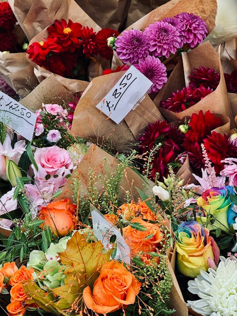 A vibrant selection of fresh flower bouquets arranged with a variety of blooms including red dahlias, purple chrysanthemums, pink roses, orange roses, and rainbow-colored roses, wrapped in brown craft