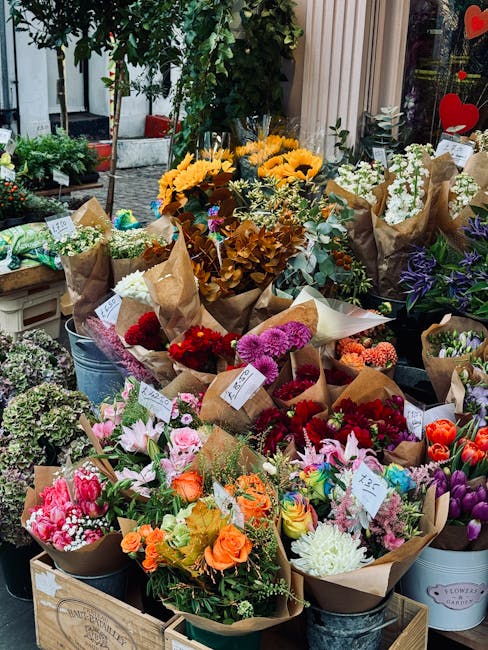An outdoor display of a vibrant assortment of fresh flower bouquets arranged in metal buckets and paper-wrapped bundles. The bouquets include a variety of colorful flowers such as bright yellow sunflo